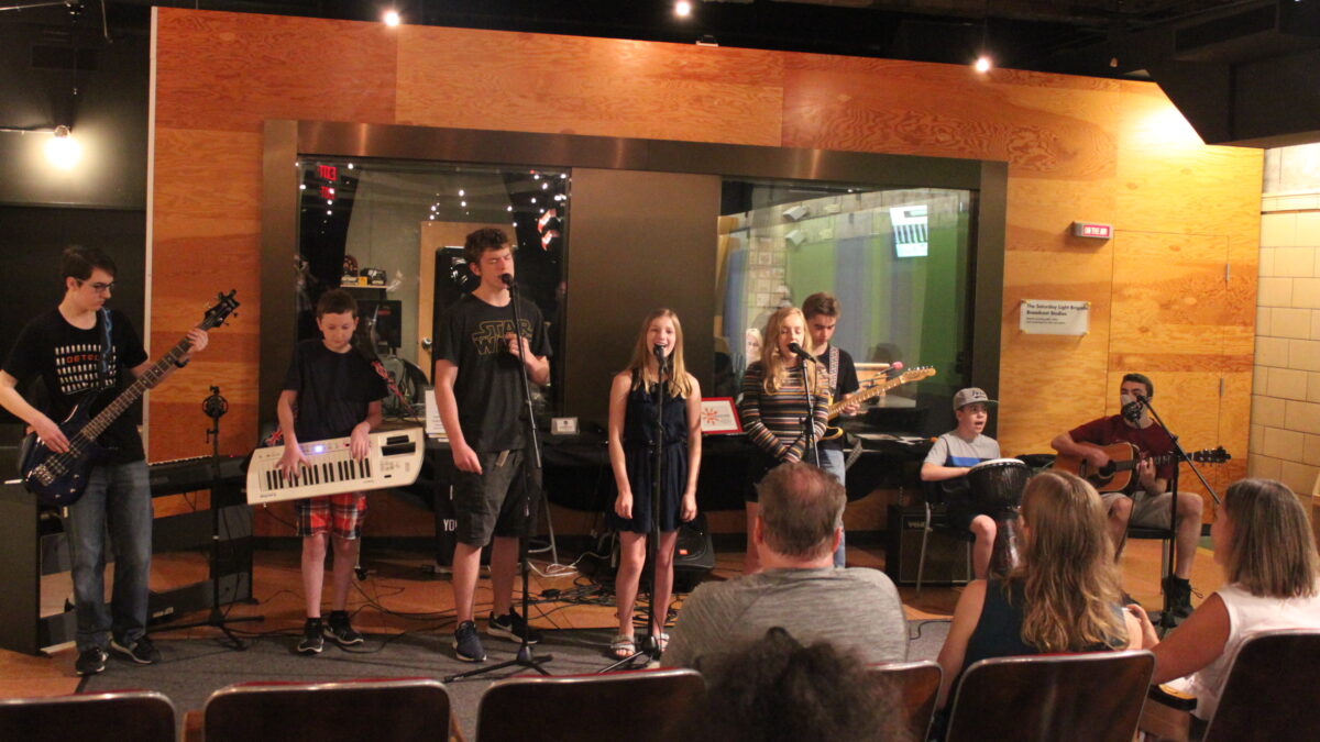 A teen playing electric bass, a youth playing keytar, three youth singing into microphones, two teens playing guitars, and a youth playing a djembe, with an audience of adults listening