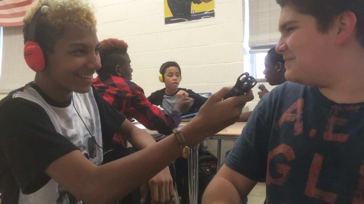 A youth wearing headphones holding a portable recorder up to another youth in a classroom