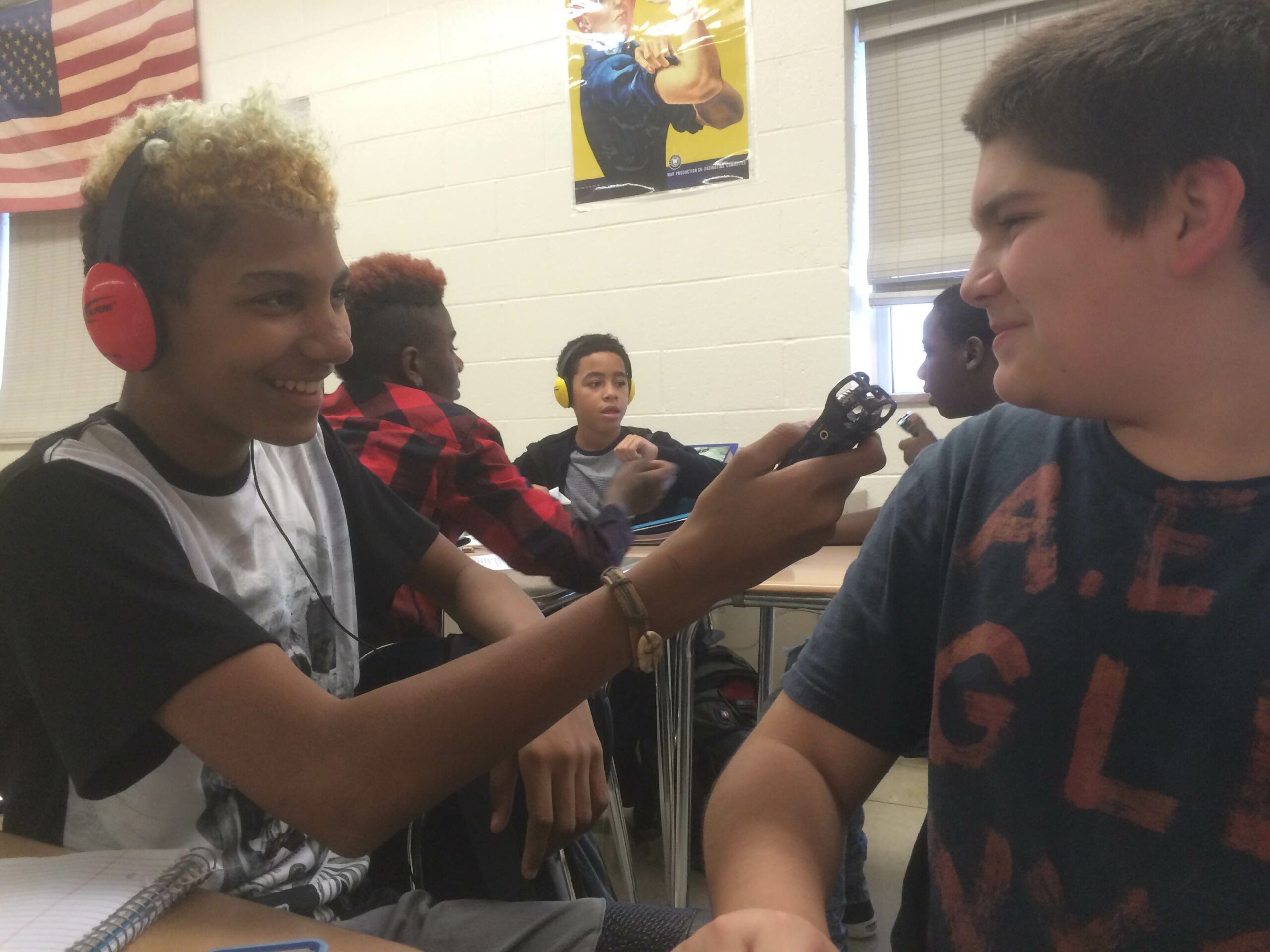 A youth wearing headphones holding a portable recorder up to another youth in a classroom