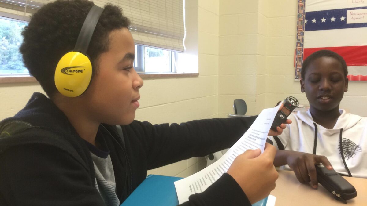 A youth wearing headphones holding a piece of paper and a portable recorder up to another youth holding a portable recorder case in a classroom