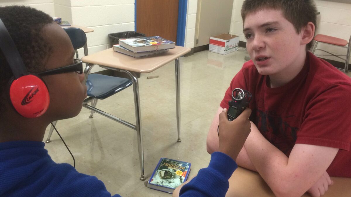 A youth wearing headphones holding a portable recorder up to another youth sitting in a classroom
