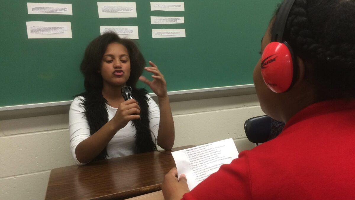 A youth speaking into a portable microphone and a youth wearing headphones holding a piece of paper in a classroom