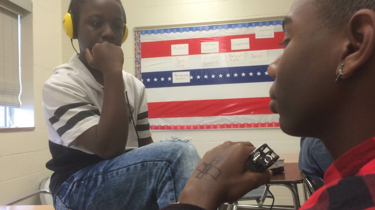 A youth holding a portable recorder and a youth wearing headphones sitting in a classroom
