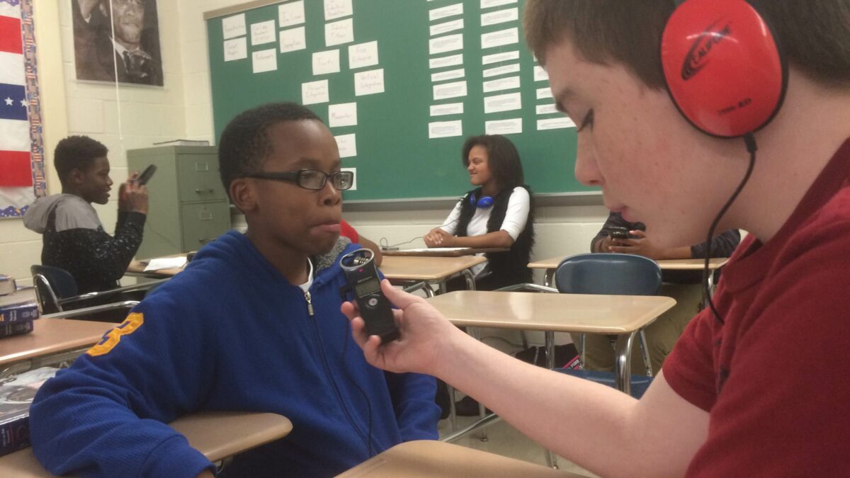 A youth wearing headphones looking down at a piece of paper and holding a portable recorder up to another youth in a classroom