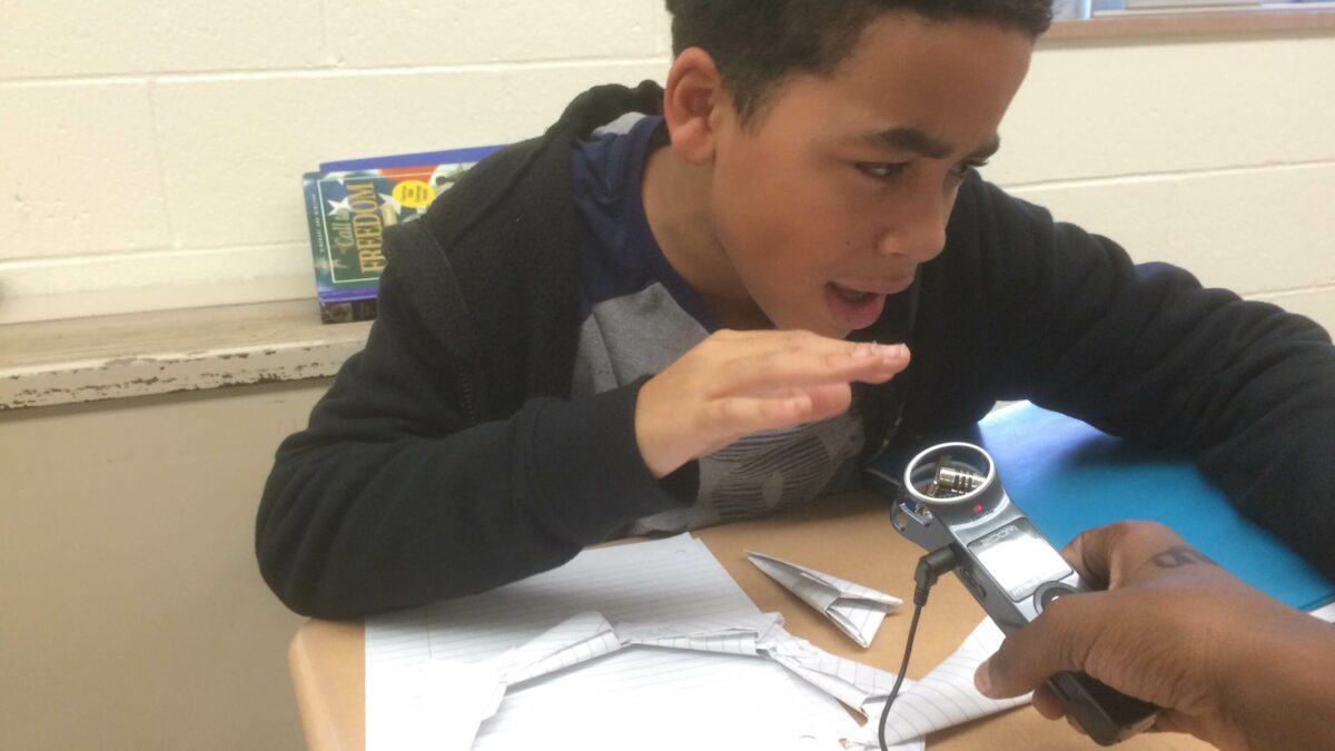 A youth talking into a portable recorder sitting at a classroom desk