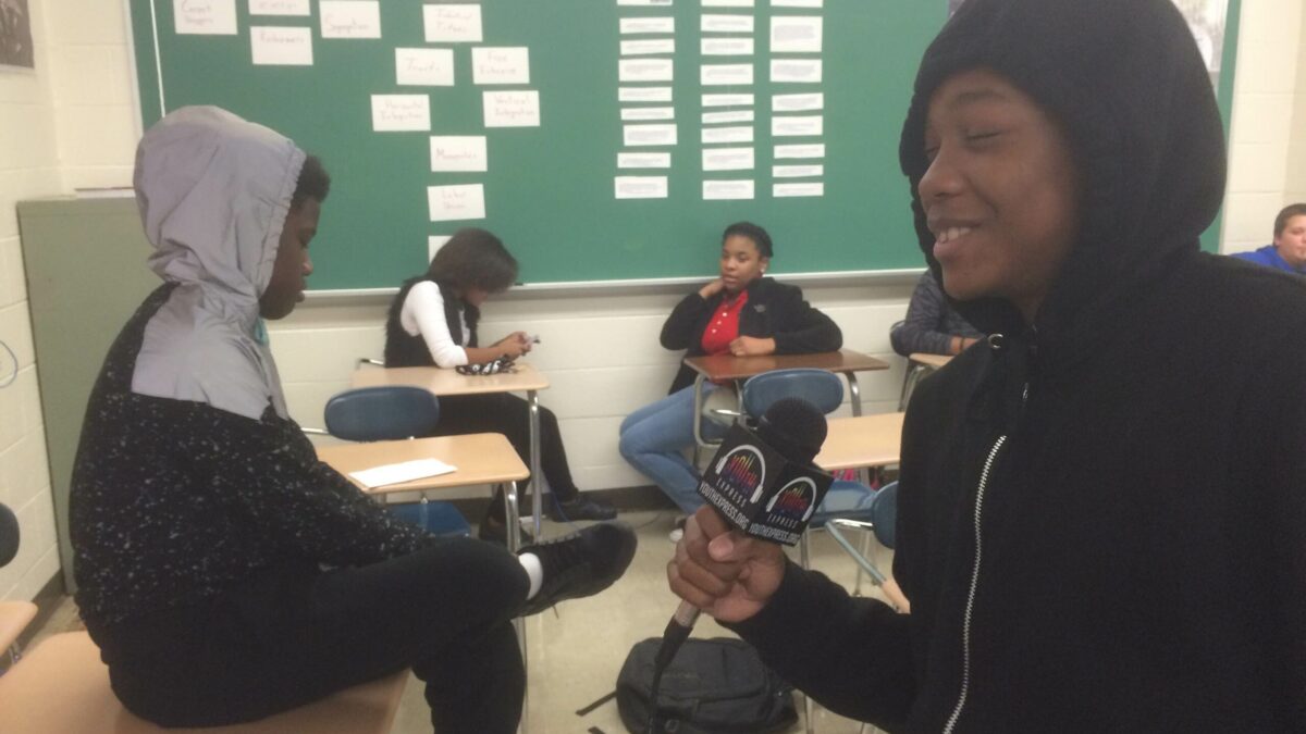 A youth smiling while holding a microphone while three youth sit in the background in a classroom