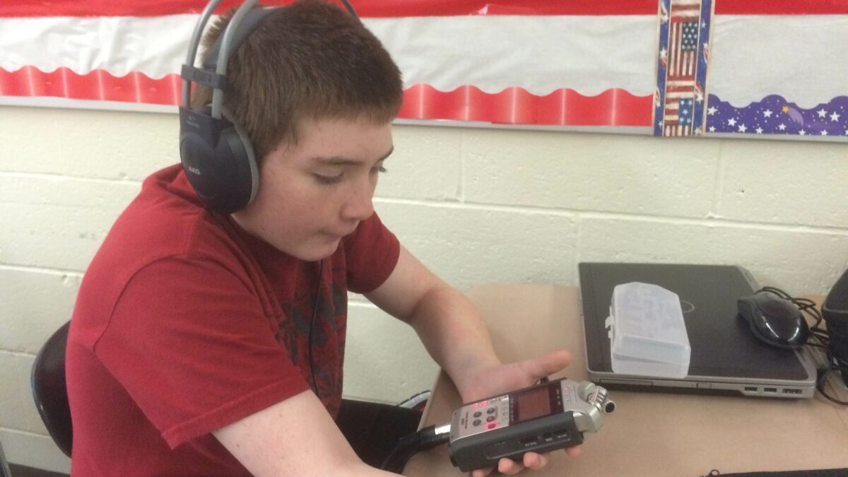 A youth wearing headphones holding a portable recorder sitting at a classroom desk