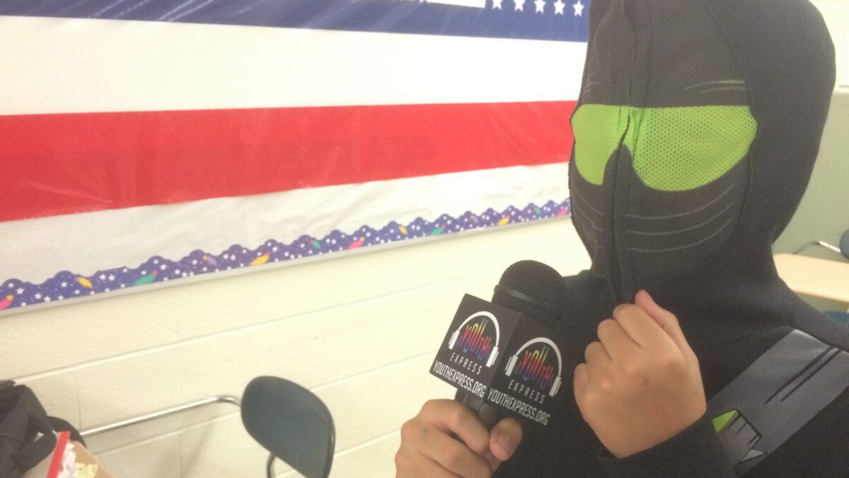 A youth wearing a hoodie and a ski mask holding a microphone in a classroom