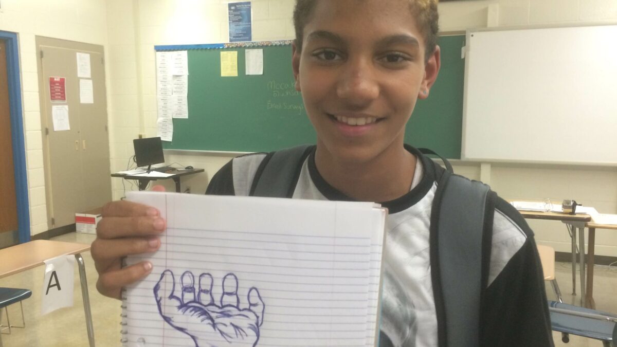 A youth holding up a drawing of a hand on a piece of notebook paper in a classroom