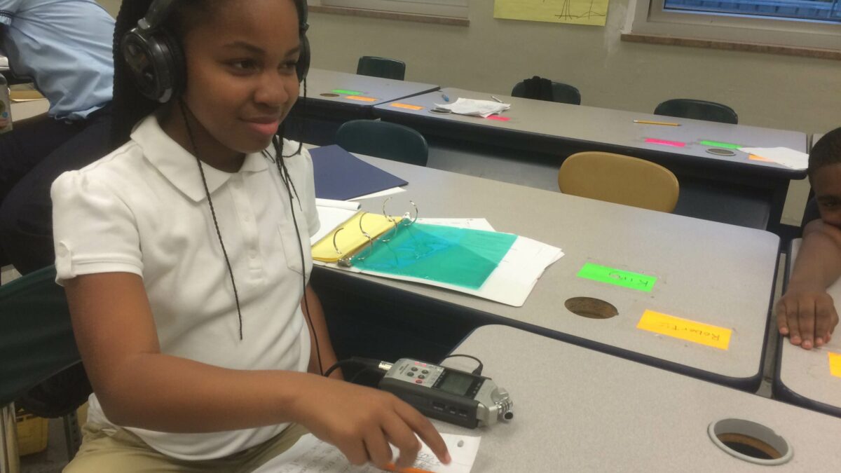 A youth wearing headphones connected to a portable recorder pointing at a piece of paper in a classroom