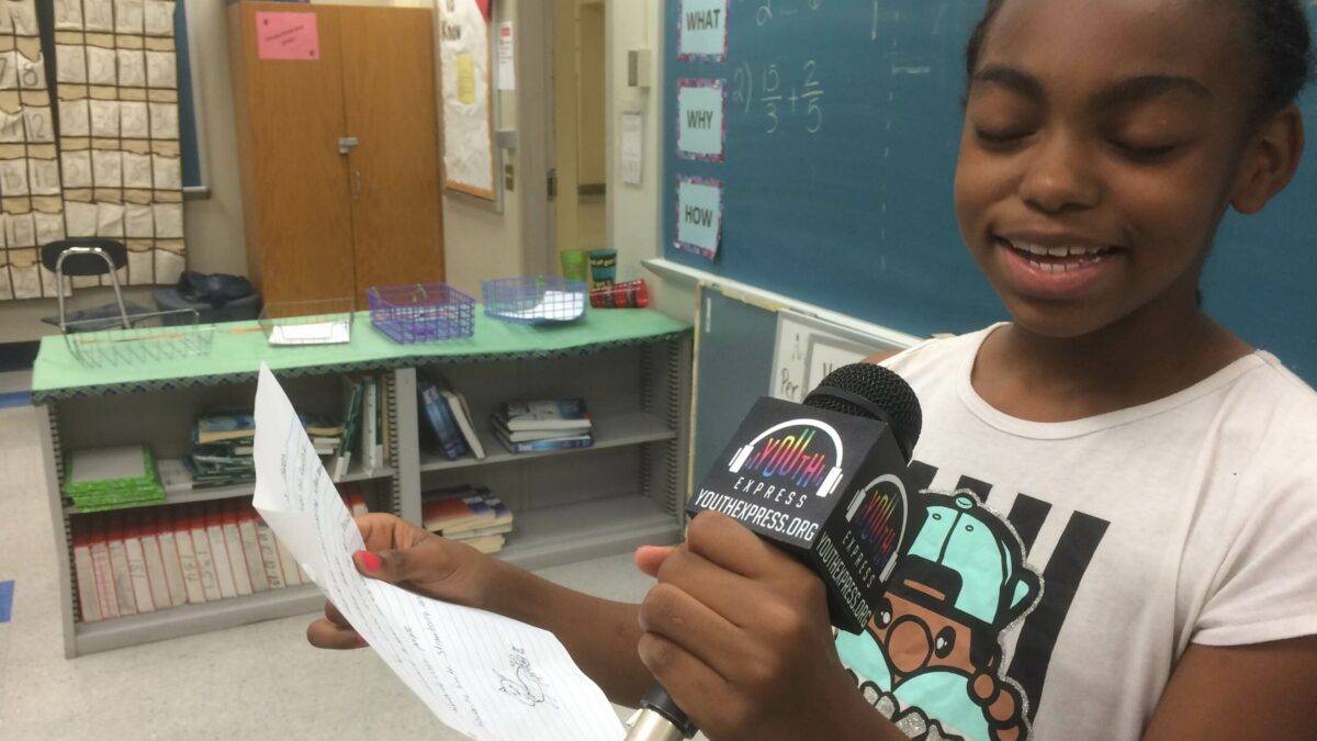 A youth holding a piece of paper speaking into a microphone in a classroom