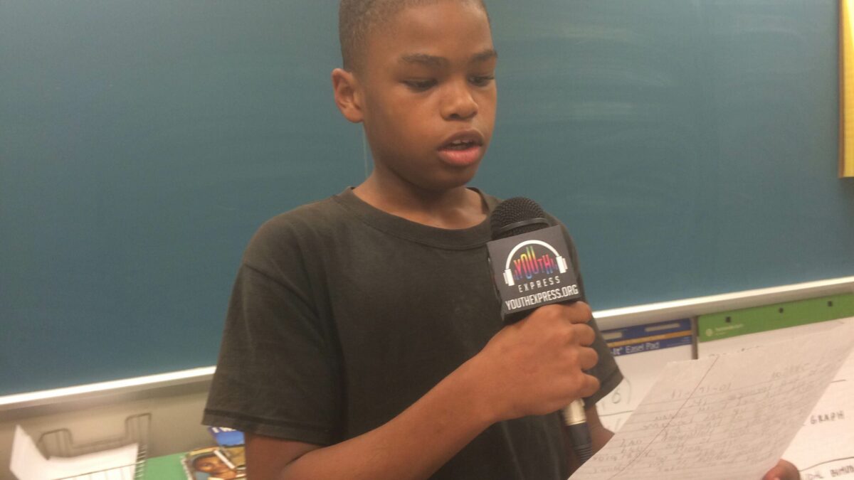 A youth holding a piece of paper speaking into a microphone in a classroom