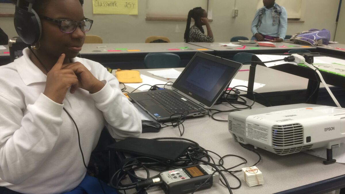 A youth wearing headphones connected to a portable recorder sitting behind a laptop computer and a projector in a classroom