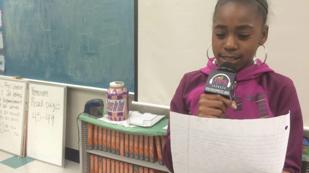 A youth holding a piece of paper speaking into a microphone in a classroom