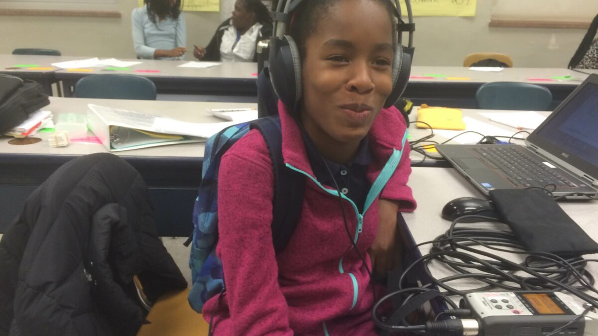 A youth wearing headphones connected to a portable recorder sitting behind a laptop computer in a classroom