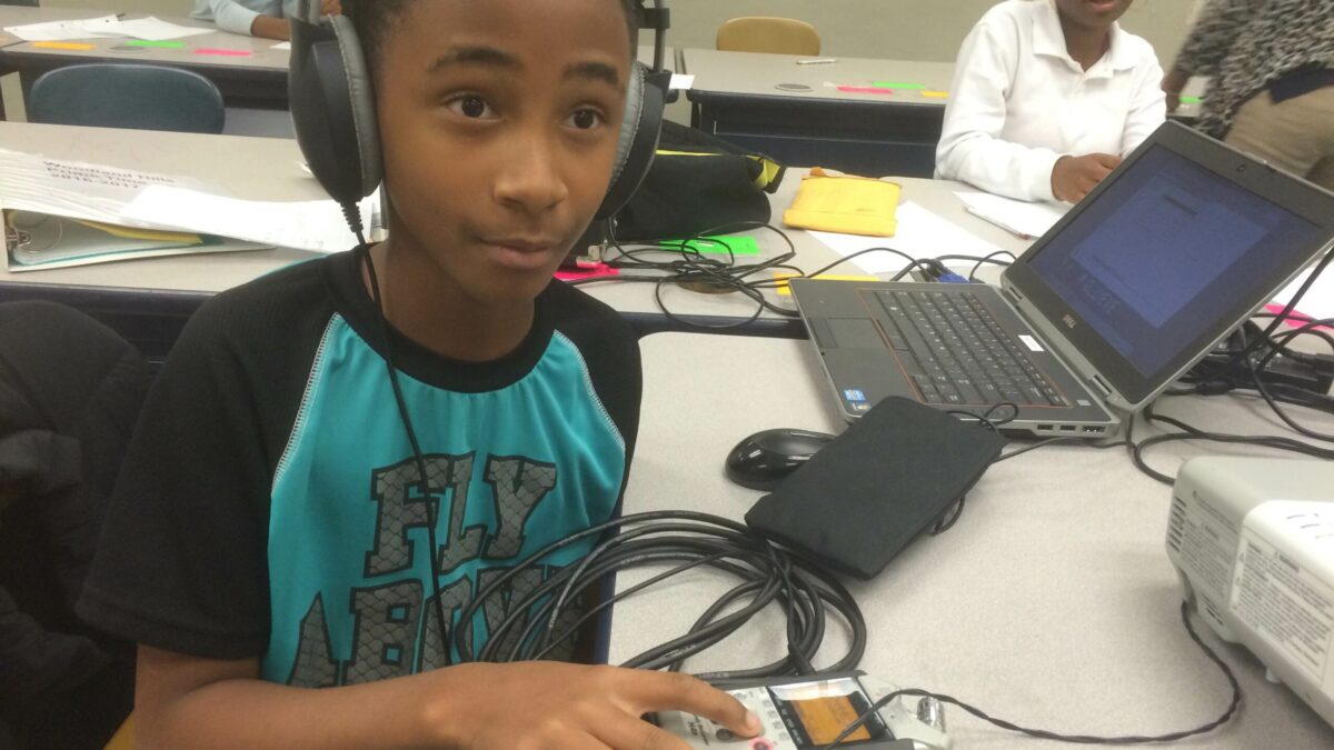 A youth wearing headphones connected to a portable recorder sitting behind a laptop computer in a classroom