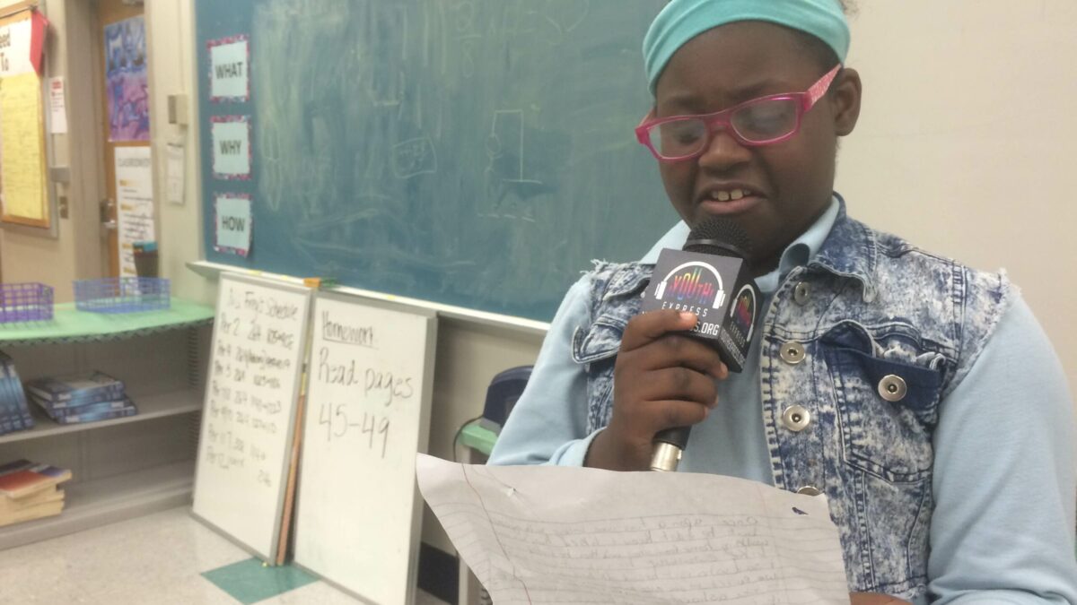 A youth holding a piece of paper speaking into a microphone in a classroom