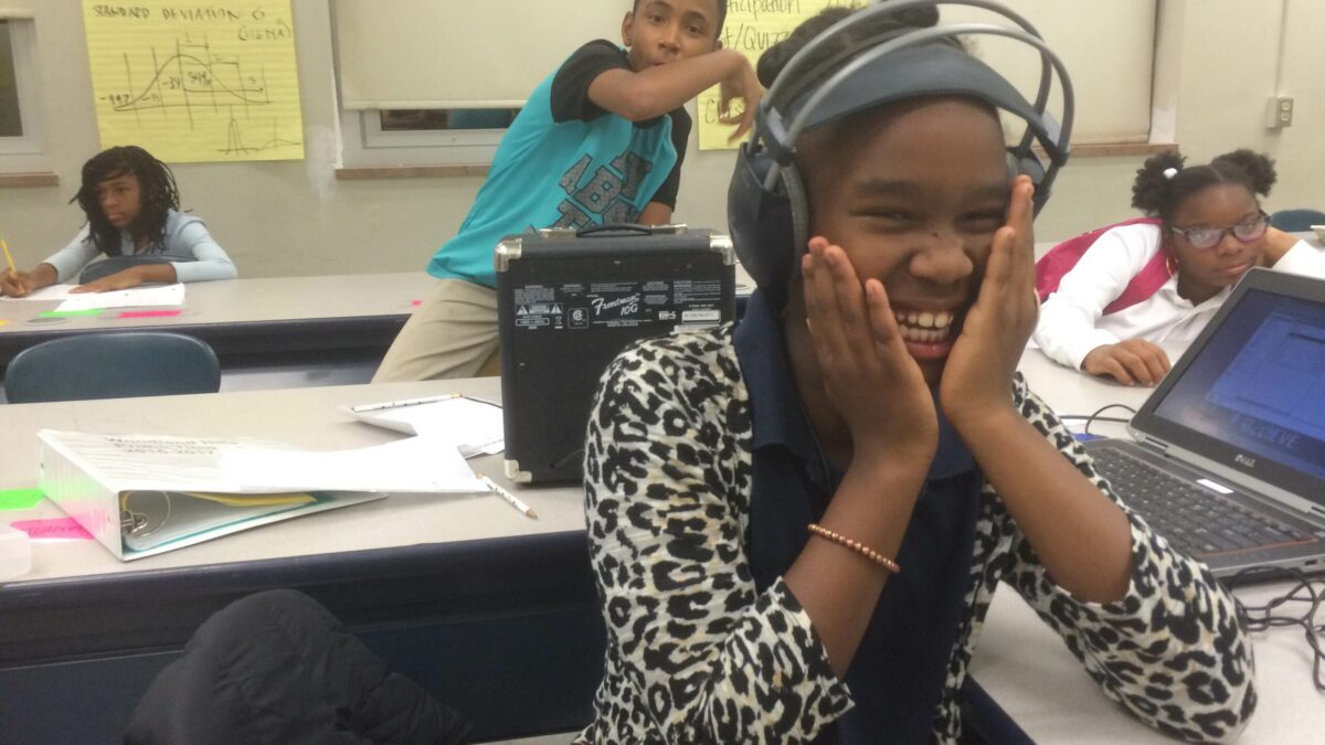 A youth wearing headphones smiling sitting behind a laptop computer in a classroom