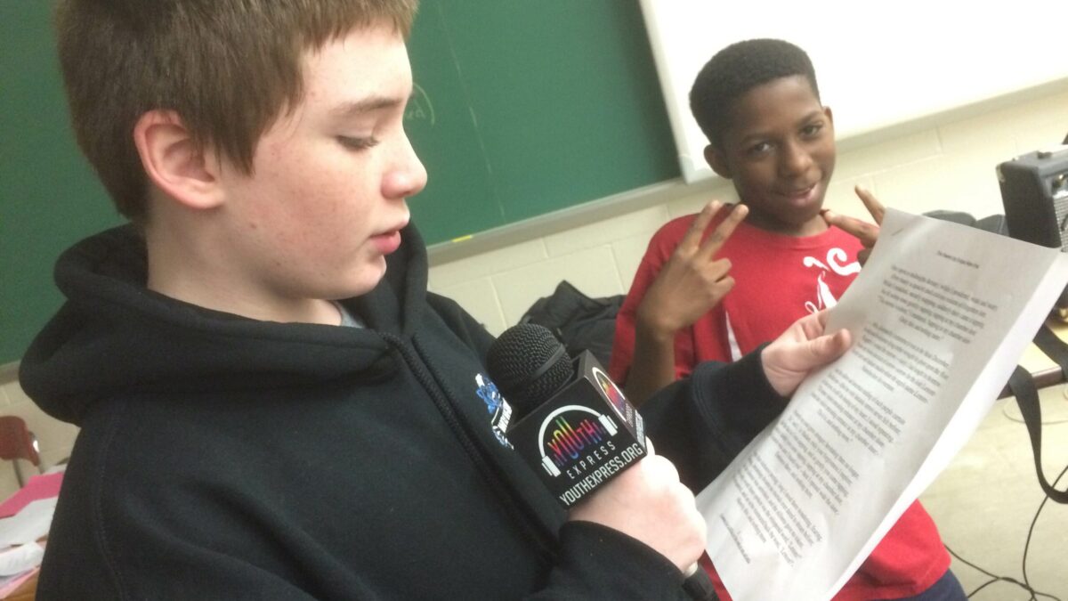 A youth holding a microphone reading a piece of paper and a youth posing for the camera making two peace signs with their hands.