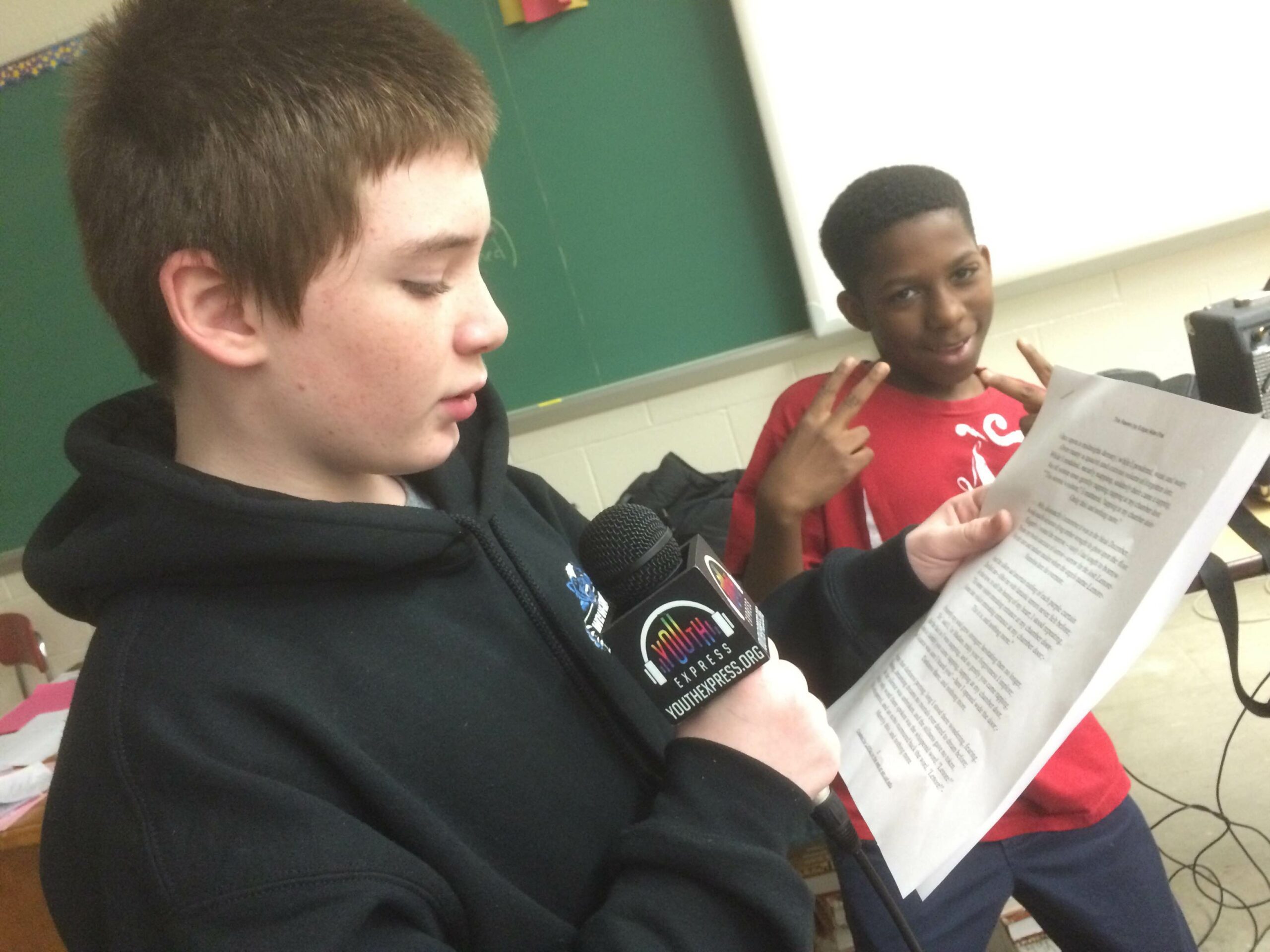A youth holding a microphone reading a piece of paper and a youth posing for the camera making two peace signs with their hands.