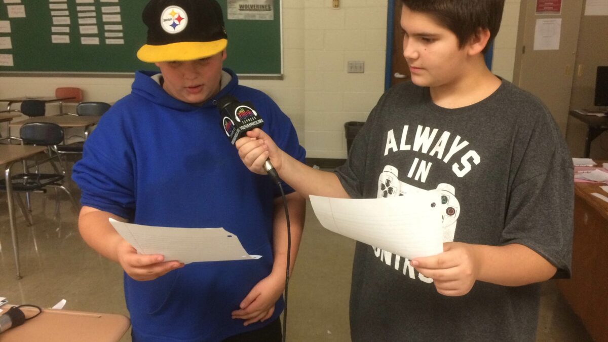 Two youth holding pieces of paper, one holding a microphone up to the other
