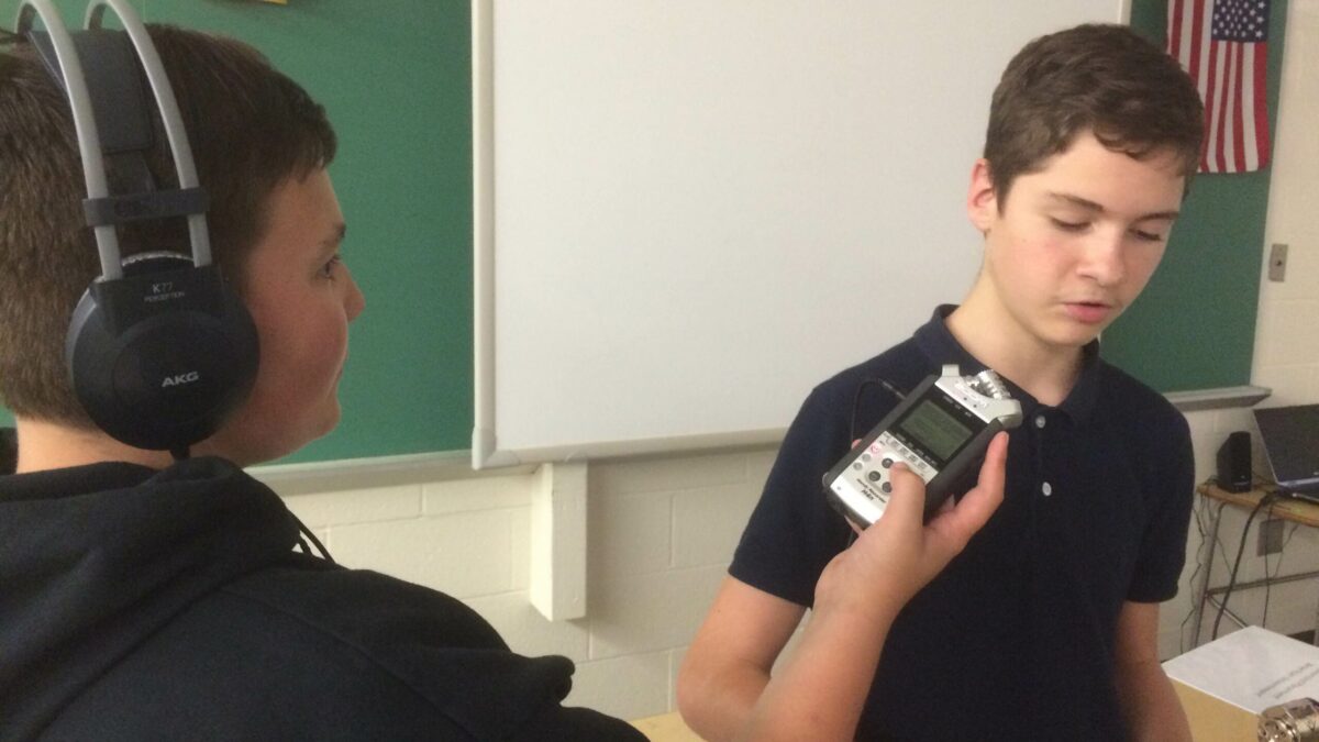 A youth wearing headphones holding up a portable recorder to another youth in a classroom