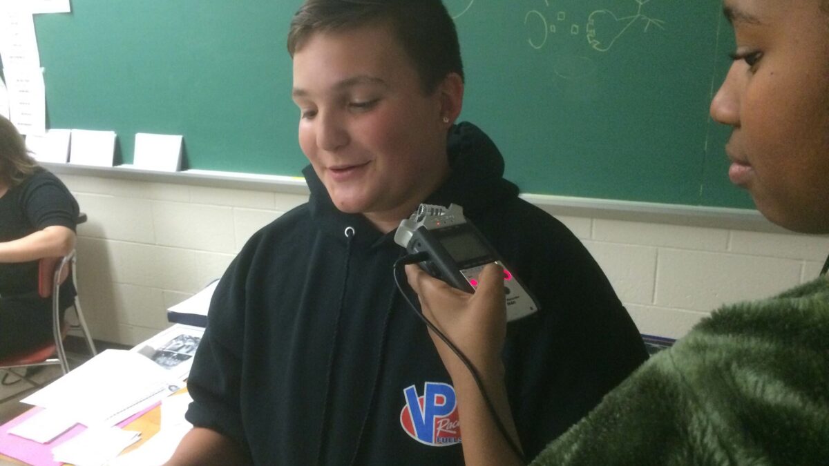 A youth holding up a portable recorder for another youth in a classroom