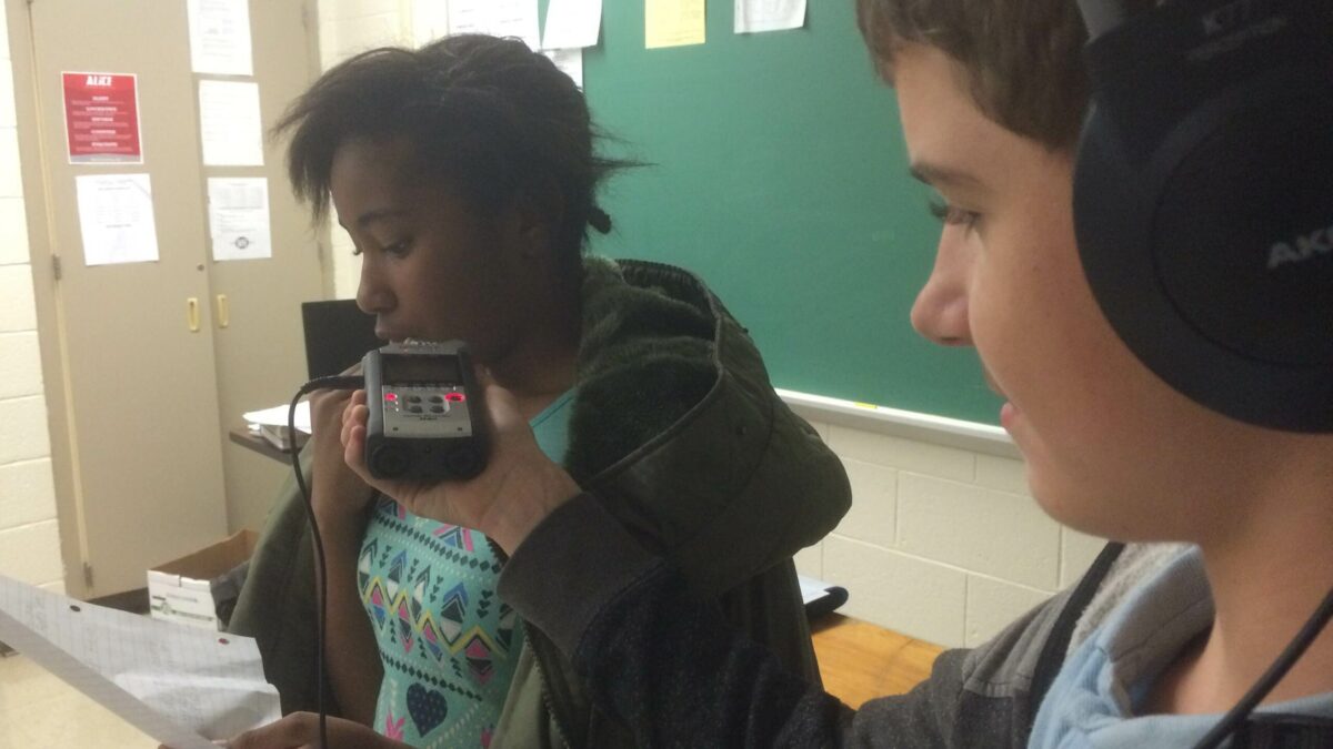A youth wearing headphones holding up a portable recorder for a youth reading from a piece of paper in a classroom