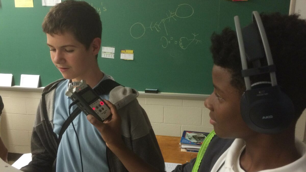 A youth wearing headphones holding up a portable recorder for a youth reading a piece of paper in a classroom