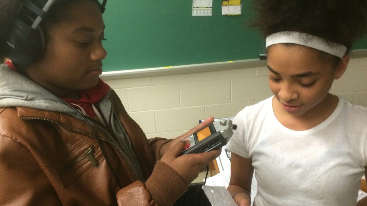 A youth wearing headphones interacting with a portable recorder and a youth holding a piece of paper in a classroom