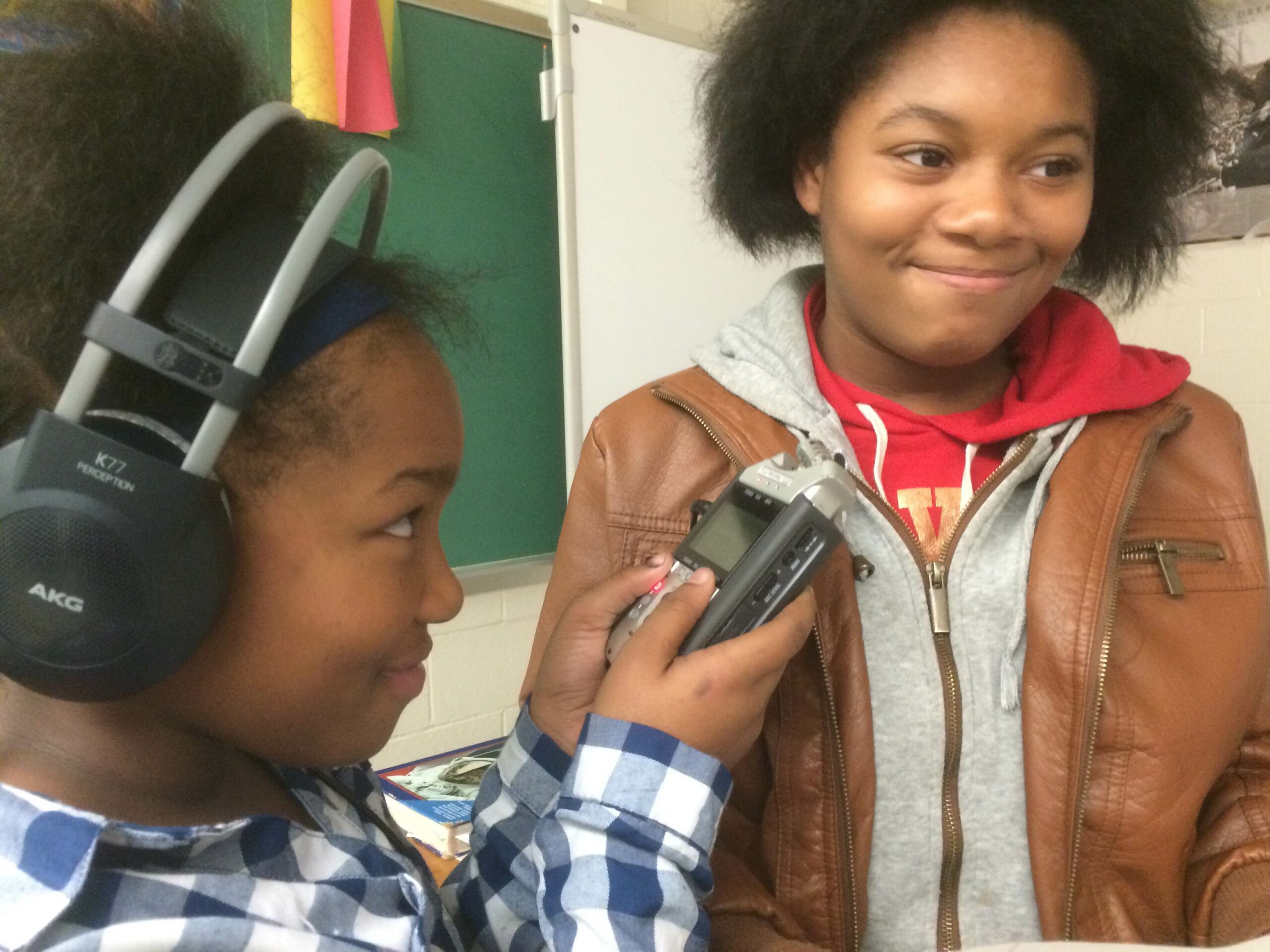 A youth wearing headphones holding up a portable recorder to another youth in a classroom
