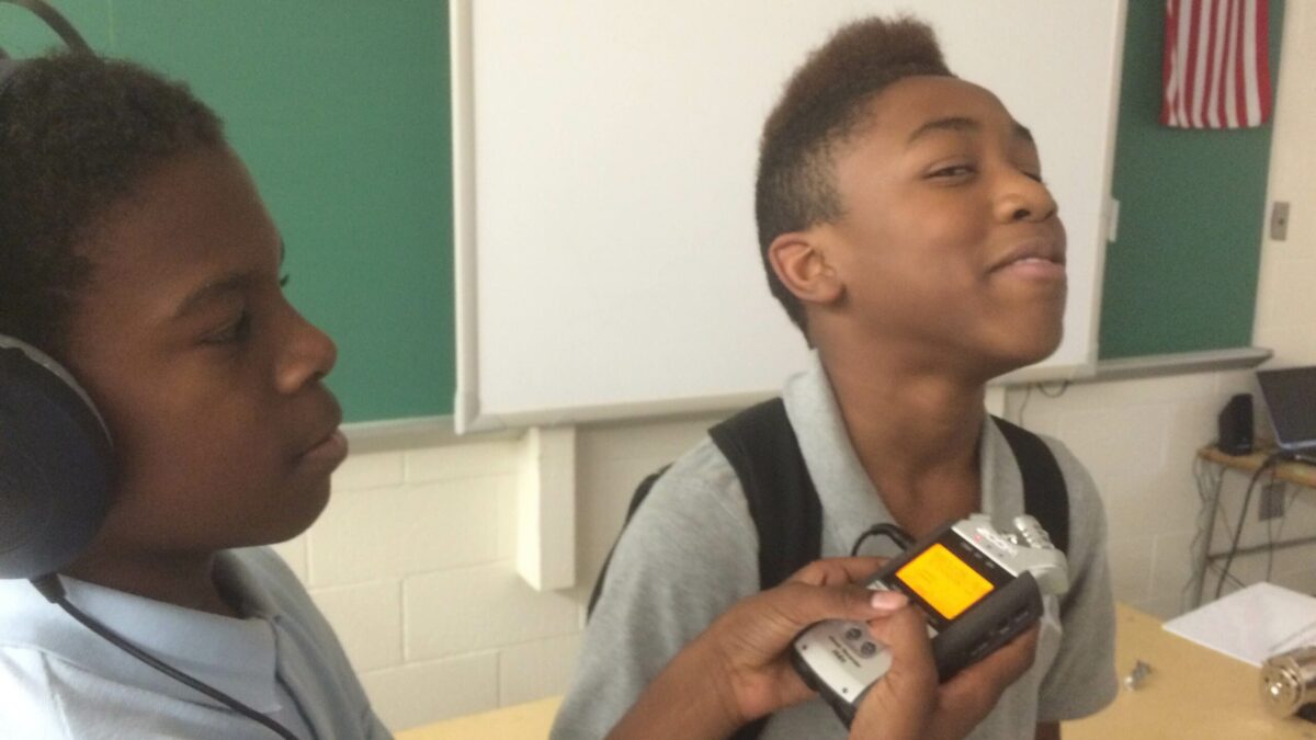 A youth wearing headphones holding a portable recorder for another youth in a classroom