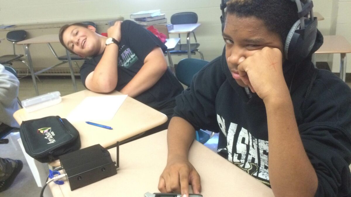 Two youth sitting at classroom desks, one wearing headphones and interacting with a portable recorder