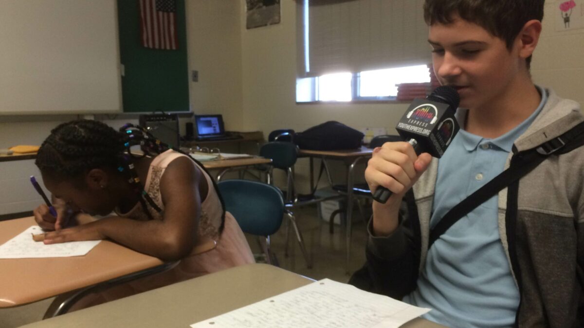 A youth writing on a piece of paper and a youth holding and speaking into a microphone while looking down at a piece of paper, both sitting at classroom desks