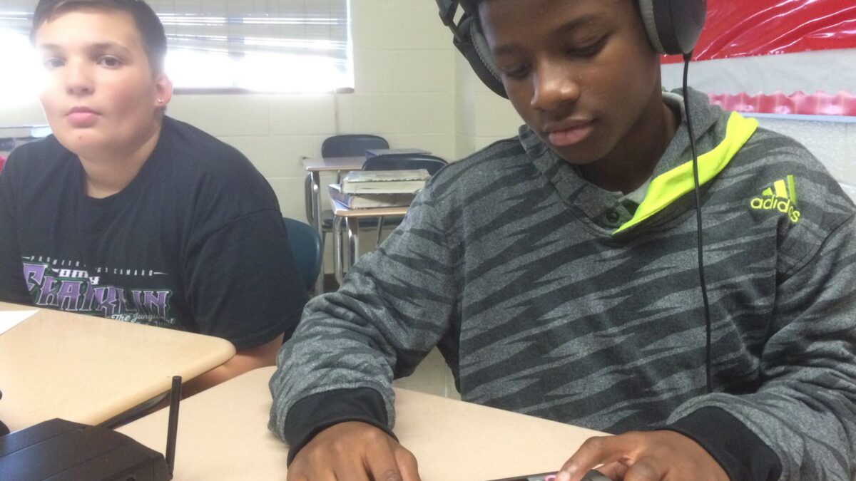 Two youth sitting at classroom desks, one wearing headphones and interacting with a portable recorder