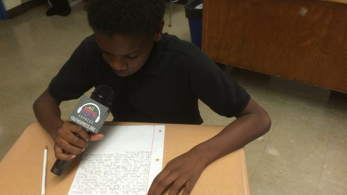 A youth holding a microphone and looking down at a piece of paper on a classroom desk