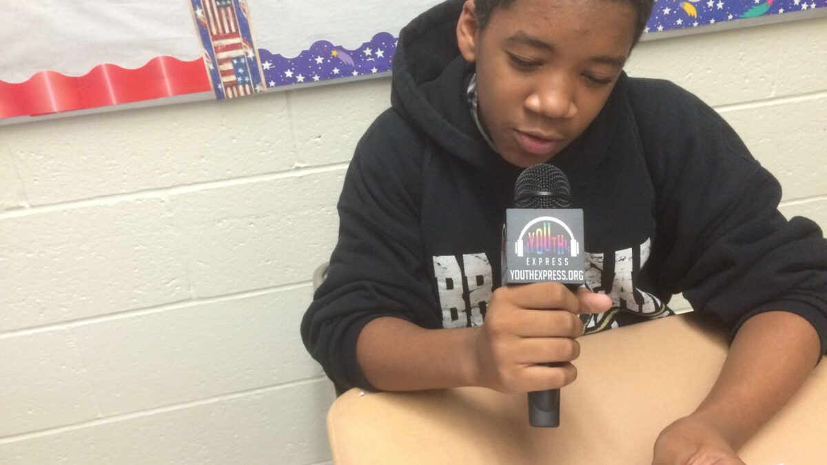 A youth holding and speaking into a microphone reading from a piece of paper in a classroom