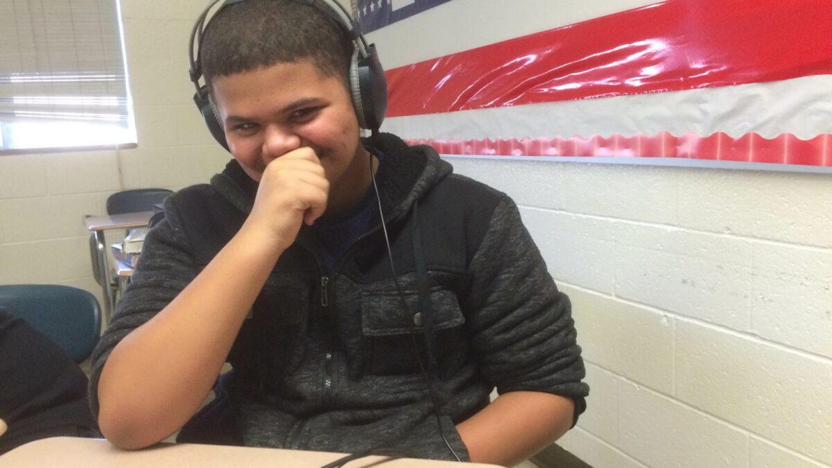 A youth wearing headphones connected to a portable recorder laughing while sitting at a classroom desk