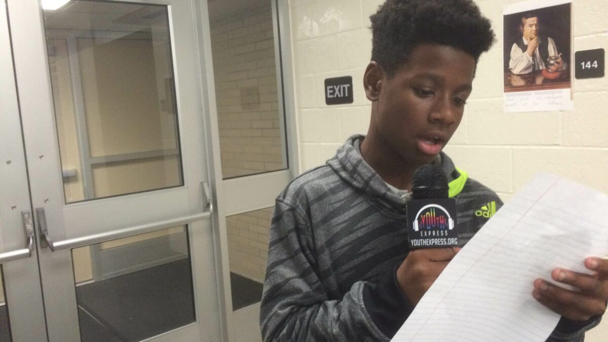 A youth holding a piece of paper and speaking into a microphone in a classroom