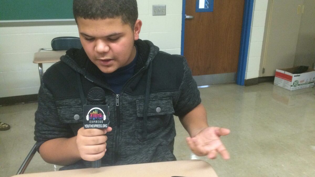 A youth sitting at a classroom desk holding and speaking into a microphone