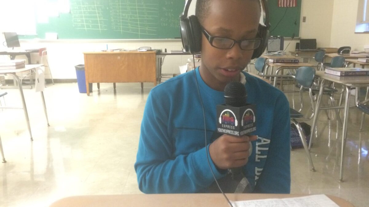 A youth wearing headphones holding and speaking into a microphone in a classroom