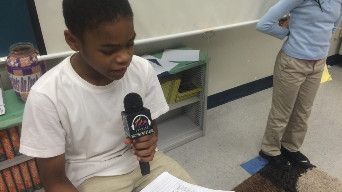 A youth speaking into a microphone looking down at a piece of paper in a classroom
