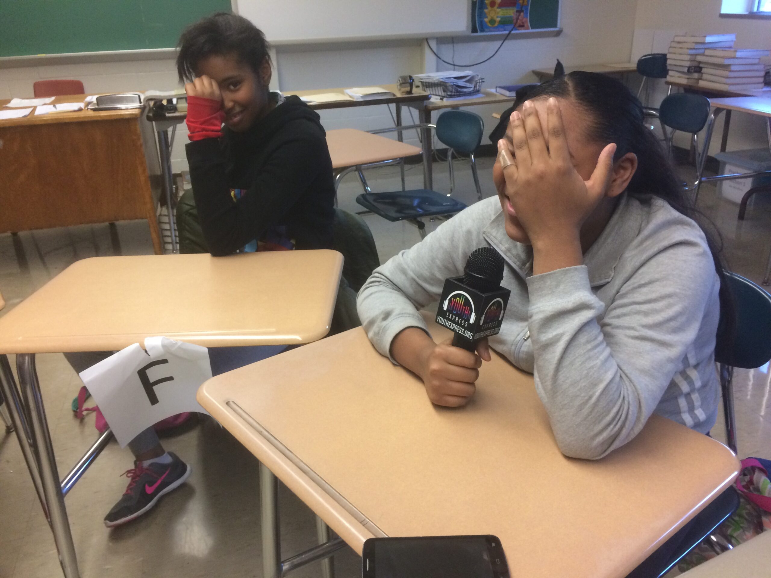 Two youth holding their faces in their hands sitting at classroom desks, one holding a microphone