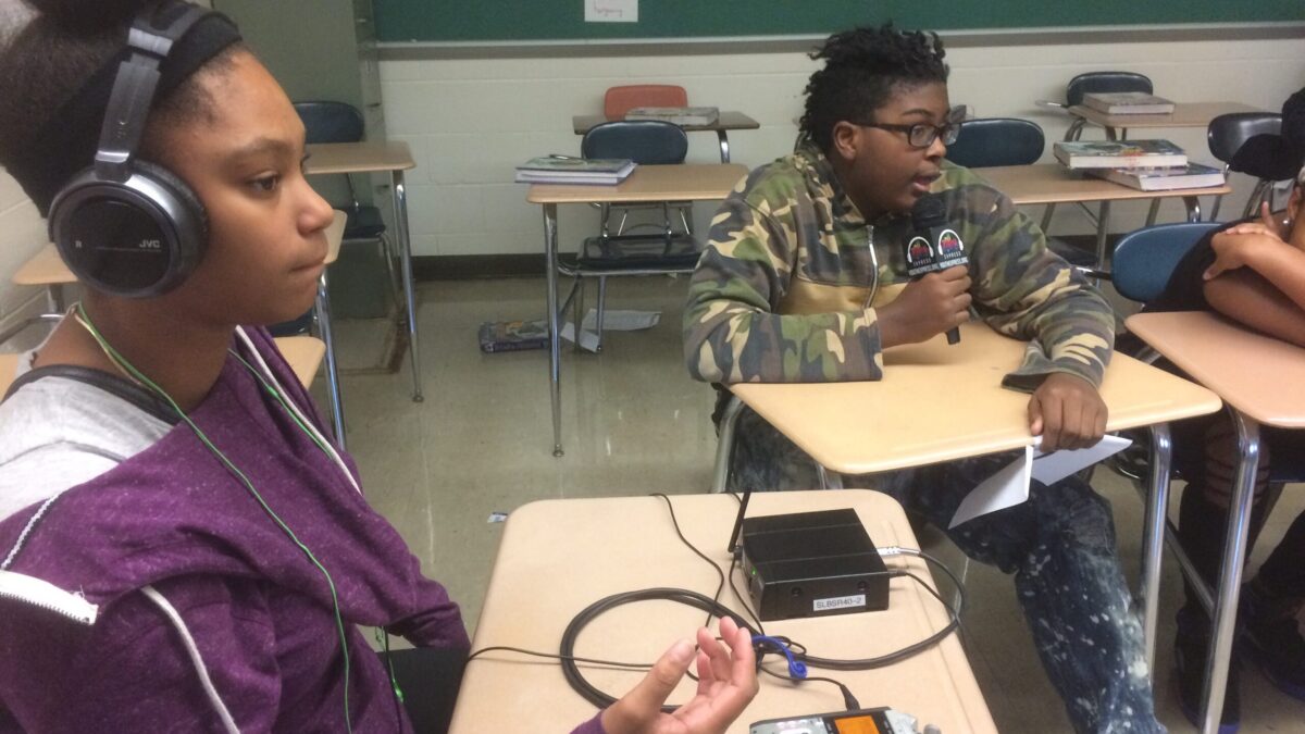 A youth speaking into a microphone connected to a portable recorder and a youth wearing headphones listening in a classroom