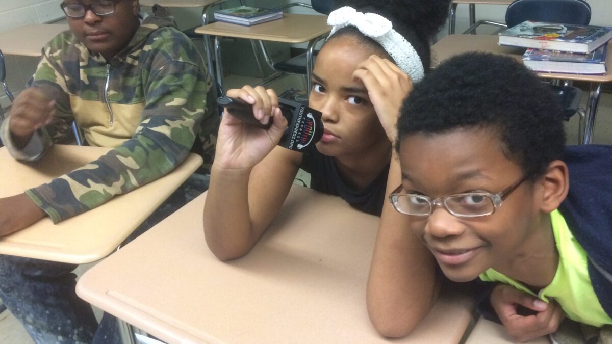Three youth sitting at classroom desks one holding a microphone
