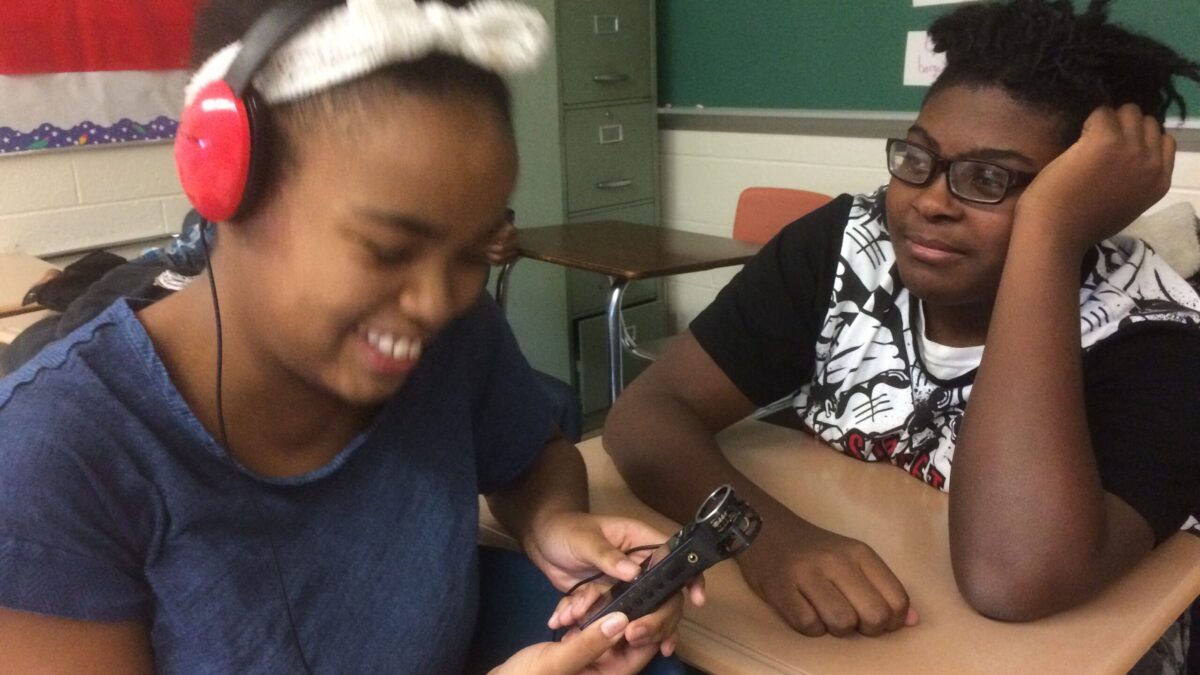 A youth sitting at a classroom desk looking at another youth wearing headphones smiling while holding a portable recorder
