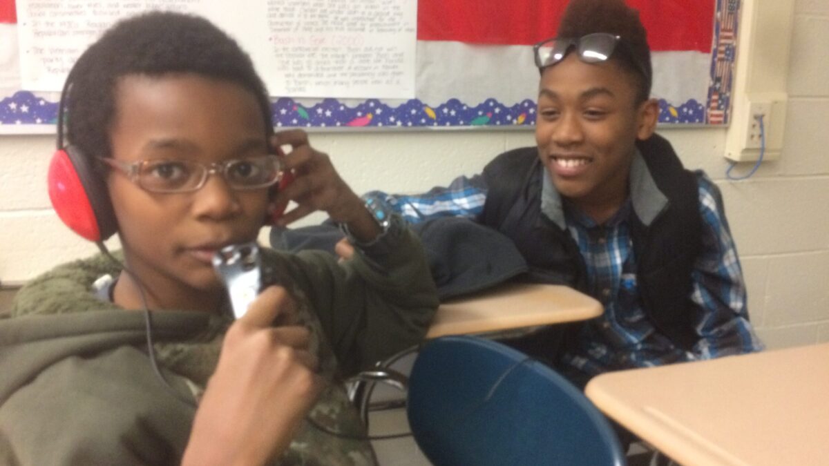 A youth smiling looking at another youth wearing headphones speaking into a portable recorder