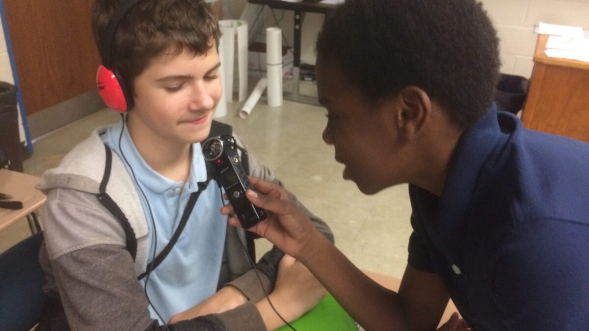 A youth holding a portable recorder up to another youth wearing headphones