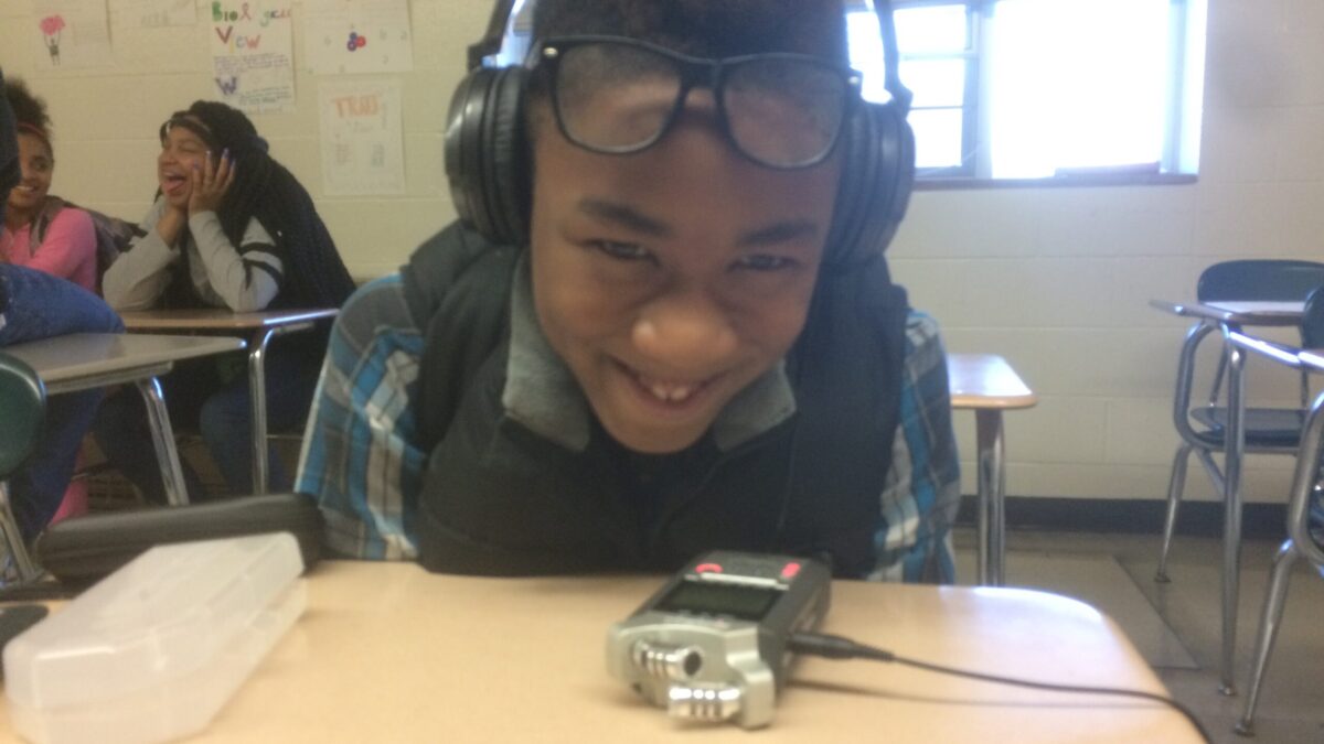 A youth wearing headphones smiling and sitting behind a portable recorder on a classroom desk.