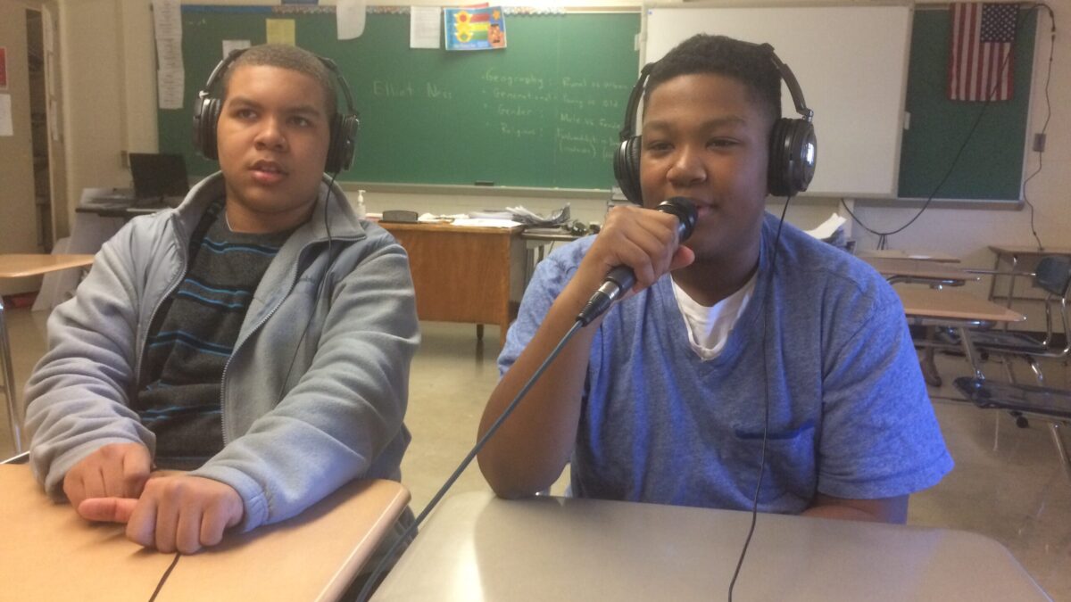 Two youth wearing headphones sitting in a classroom, one holding and speaking into a microphone